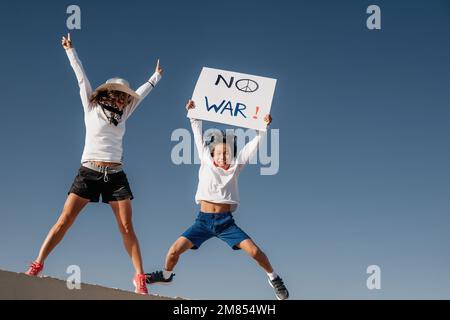 Una madre caucasica in un cappello bianco, salta accanto al suo figlio asiatico tenendo un segno che dice 'nessuna guerra'. concetto di guerra e pace. conflitto e protesta. subacquei Foto Stock