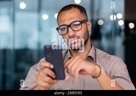 Serio uomo d'affari pensante primo piano, uomo in possesso di smartphone e la lettura di notizie online, lavoratore all'interno di ufficio vicino alla finestra in camicia e occhiali utilizzando il telefono digitando messaggio. Foto Stock