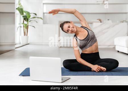 Giovane donna in forma in abbigliamento sportivo pratica yoga a casa con formazione online sul notebook, guardando lezioni virtuali e tutorial. Quarantena sport, fitness e allenamento. Il coach conduce una lezione di yoga virtuale Foto Stock