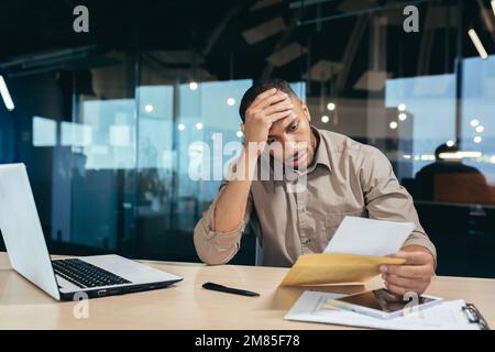 Uomo d'affari pensivo all'interno dell'ufficio che legge la lettera di notifica, l'uomo ha ricevuto la busta che pensa alla decisione che siede al tavolo ed al laptop all'interno dell'ufficio, messaggio difettoso di notizie. Foto Stock