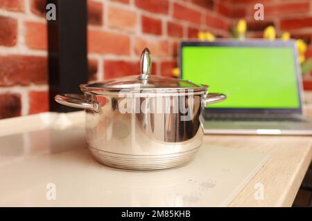 Pentola in acciaio su elegante stufa a induzione bianca e computer portatile con schermo verde con chiave cromata in cucina su sfondo muro di mattoni. Cucina e vlogging concetto. Foto Stock