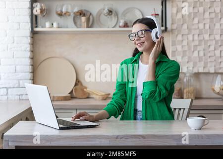 Giovane donna bella seduta in cuffie a casa al tavolo da cucina e utilizzando il notebook, guardare online webinar, film, studio, ascoltare musica. Foto Stock