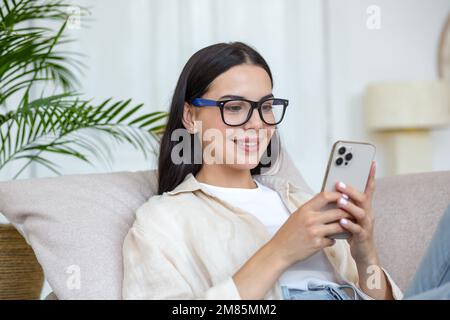 Una giovane bella donna è sdraiata sul divano di casa con un telefono in mano. Effettua ordini di cibo per la casa, shopping in un negozio online. Foto Stock