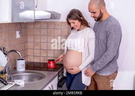 Una giovane donna incinta che fa il caffè in cucina, con il marito che la sostiene. Foto Stock