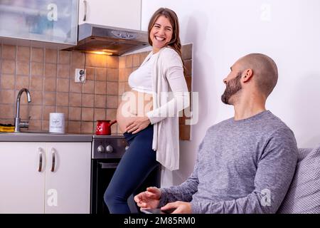 Una giovane donna incinta che fa il caffè in cucina, mentre suo marito si rilassa sul divano, lavorando su un computer portatile. Foto Stock