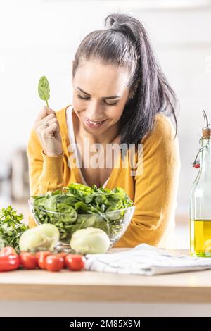 Bella giovane donna che tiene una foglia di spinaci, sorridente, verdure fresche sul tavolo accanto a lei. Foto Stock