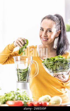 Donna sorridente che prepara il frullato di spinaci, mettendo le foglie nel frullatore. Concetto di stile di vita sano e mangiare. Foto Stock