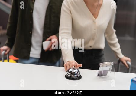 vista parziale della donna che suona il campanello alla reception vicino a un ragazzo sfocato con smartphone in hotel, immagine stock Foto Stock