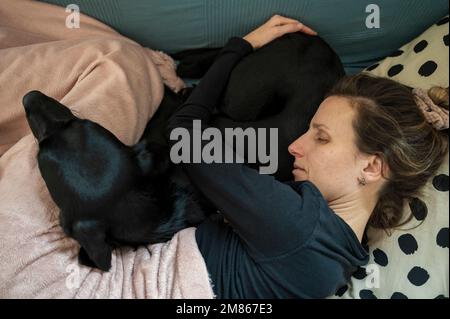 Giovane donna malata con un'influenza sdraiata su un divano con il suo cane nero labrador Retriever coccolato accanto a lei. Foto Stock