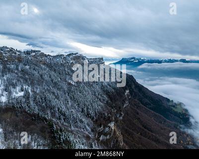 colpo con un drone dalla cima di una montagna, che offre una vista dei pini adornati da una polvere di neve sotto un cielo disseminato di nuvole intermittenti Foto Stock