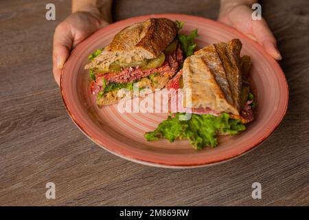 Mani di un piatto di presa umana di un panino fresco appetitoso sul primo piano del tavolo, vista dall'alto. Servizio di hamburger di carne a fette con verdure e verdure Foto Stock