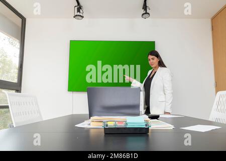 una giovane donna dirigente punta a uno schermo verde, dove sta facendo una presentazione online in una sala riunioni. Foto Stock