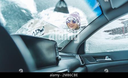 Giovane donna sorridente che pulisce la prima neve dal parabrezza della sua auto. Vista dall'interno dell'auto con angolazione ridotta. Foto di alta qualità Foto Stock