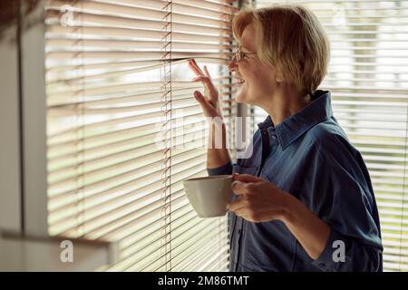 Attraente donna matura bevendo una bevanda calda, caffè o tè e guardando attraverso la finestra Foto Stock
