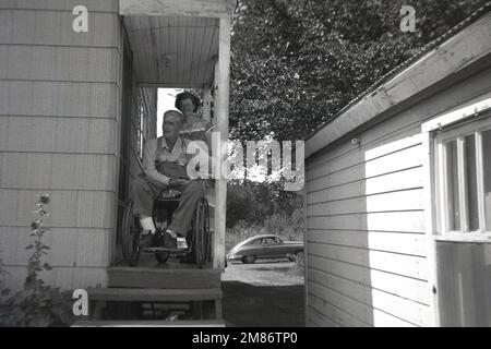 1950s, storico, centro-ovest America, un anziano seduto in una sedia a rotelle, moglie con lui, sul portico rialzato laterale e l'ingresso di una casa rurale a un piano, gradini aperti fino al portico. Un'auto americana parcheggiata all'esterno in una corsia dietro la proprietà. Foto Stock