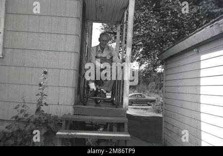 1950s, storico, centro-ovest America un anziano seduto in una sedia a rotelle, sul portico rialzato laterale e l'ingresso alla sua casa rurale a un piano, passaggio aperto fino al portico. Un'auto americana parcheggiata all'esterno. Foto Stock