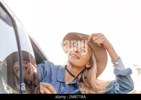 Sognando donna con gioia lasciare la città che si stacca testa fuori dal finestrino dell'auto aperto e guardare fino al cielo. Giovane donna bionda in cappello con umore felice andare in auto Foto Stock