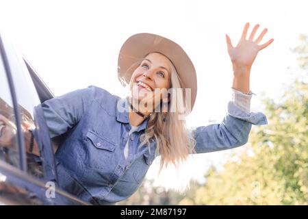 Donna divertente con gioia lasciare la città che si sta attaccando testa fuori dal finestrino dell'auto aperto e ondeggiando la mano. Giovane donna bionda in cappello con umore felice andare a viaggio in auto Foto Stock