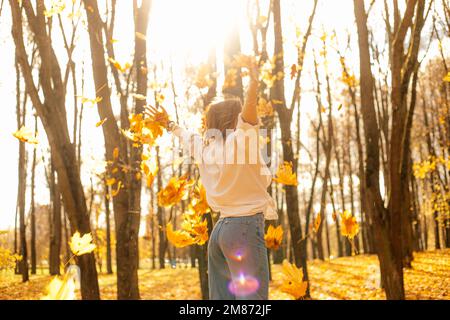 Vista posteriore di meravigliosa, donna bionda incurante camminare nella foresta dorata, raccogliere e gettare quercia e foglie di acero come pioggia Foto Stock