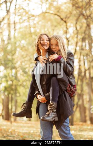 Verticale giocoso positivo, amorevole, allegra famiglia giovane madre tenendo la figlia sul retro, passeggiata nella foresta d'autunno dorato Foto Stock