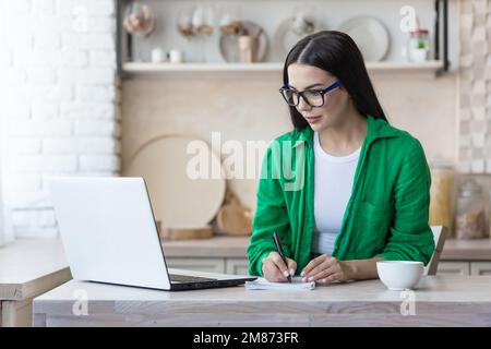 Una giovane donna seria sta avendo un colloquio di lavoro remoto in linea. Seduti a casa in cucina con un computer portatile. Parla con una videochiamata, si presenta, supera l'esame, scrive in un notebook. Foto Stock