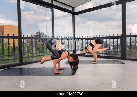 Due giovani donne davanti alle finestre dello studio di yoga, che fanno la posa di un corvo di loto mezzo. pose avanzate di yoga. Foto Stock