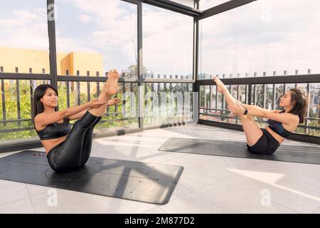 Le giovani donne che fanno la barca posa in studio yoga con finestre sullo sfondo, indossando abbigliamento sportivo. pose avanzate di yoga. Foto Stock