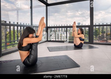 Due belle giovani donne latine che fanno pose yoga avanzate in studio, indossando abbigliamento sportivo, con finestre sullo sfondo. pose avanzate di yoga. Foto Stock