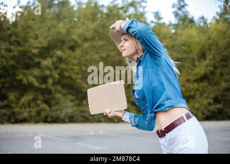 Donna bionda esausta curiosa tenere cappello hitchhiking con piastra di cartone vuota e cercare il traffico di auto da strada autostradale Foto Stock