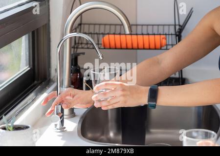 Le mani di una donna che tiene un bicchiere che si riempie di acqua dal filtro del rubinetto. Foto Stock