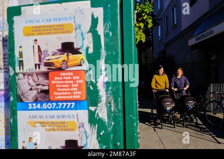 Due donne ebraiche ortodosse spingono passeggini sul marciapiede vicino a Lee Avenue con poster di servizio auto locali in foreground.Williamsburg.Brooklyn.New York City.USA Foto Stock