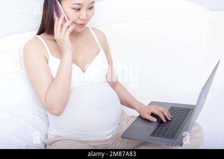 Le donne incinte si sono sedute sul letto utilizzando computer portatili e dispositivi mobili Foto Stock