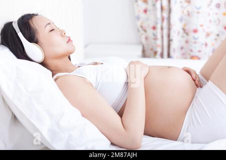 Donna incinta sdraiata a letto ascoltando musica Foto Stock