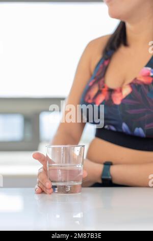 primo piano delle mani di una donna che tiene un bicchiere d'acqua, su un bancone da cucina in uno studio sportivo. Foto Stock