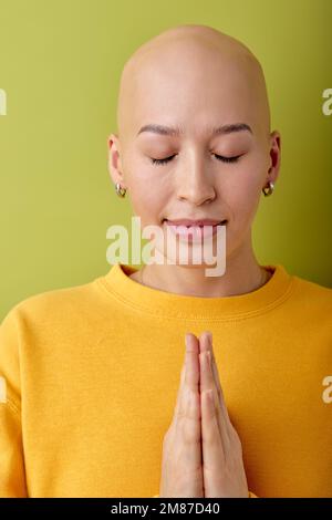 bella giovane caucasica calva senza capelli donna che prega, tenendo le mani insieme, indossare abiti casual in piedi isolato su sfondo verde, chiedendo Foto Stock