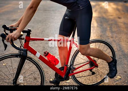 Primo piano di un giovane sportivo con gambe robuste in piedi su strada con la moto rossa. tramonto, sera. Concetto di ciclismo. uomo irriconoscibile in spo Foto Stock