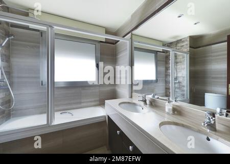 Angolo di una toilette con un grande specchio a parete, un lavabo a due ciotole in porcellana bianca e marmo beige e un'unità idromassaggio con divisorio in vetro Foto Stock