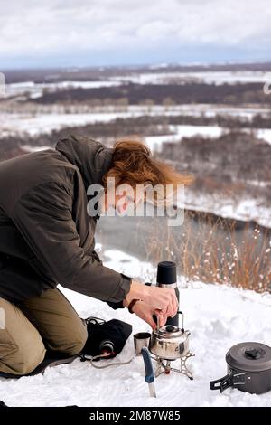 Maschio mettere piccolo bollitore turistico sul fuoco nella foresta invernale durante il campeggio, in viaggio. da solo. fiume o lago sullo sfondo, splendido paesaggio. viaggi Foto Stock