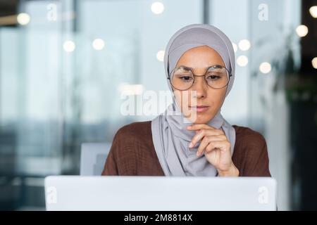 Primo piano di pensare donna d'affari in hijab in ufficio moderno, seria donna musulmana guardando lo schermo del notebook pensando alla decisione, donna sul posto di lavoro di successo. Foto Stock