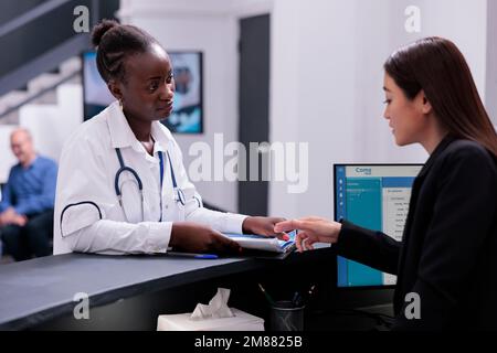 Addetto alla reception che aiuta il medico con gli appuntamenti di visita di controllo mentre lavora al banco di registrazione dell'ospedale nell'area di attesa. Medic esamina l'anamnesi dei pazienti prima della loro consultazione. Foto Stock