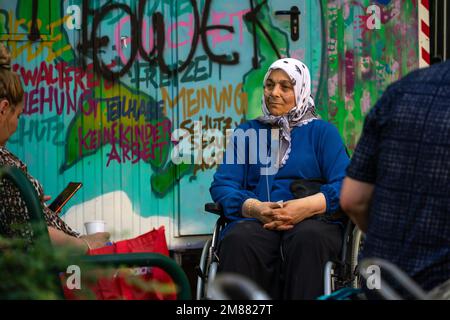 Donna anziana in una sciarpa che siede in una sedia a rotelle e guarda la sua amica, con un contenitore coperto da graffiti dietro di lei Foto Stock