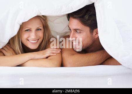 Accoppiare sotto la copertura. Una coppia sdraiata su un letto sotto il piumino e sorridente. Foto Stock
