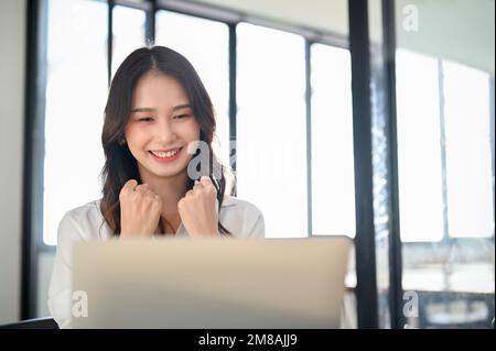 Giovane lavoratrice asiatica che guarda lo schermo del notebook, festeggia le sue buone notizie, felice del risultato del suo progetto. Foto Stock