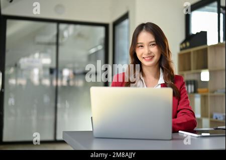 Affascinante donna d'affari asiatica millenaria o manager femminile in vestito rosso siede alla sua scrivania, sorridendo e guardando la fotocamera. Foto Stock