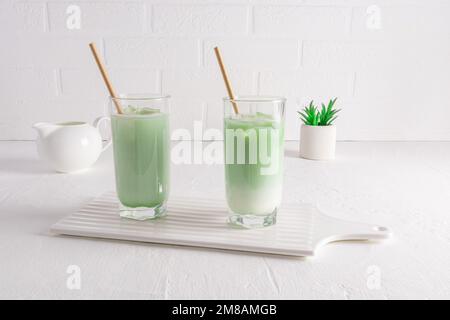 una sana lattina di matta con latte in bicchieri alti con paglia su una tavola di ceramica bianca contro una parete bianca. bevanda biologica naturale Foto Stock