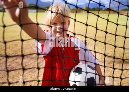 Il suo obiettivo è in vista. Una bambina che tiene una sfera di calcio mentre guarda attraverso la rete di obiettivo su un campo di calcio. Foto Stock
