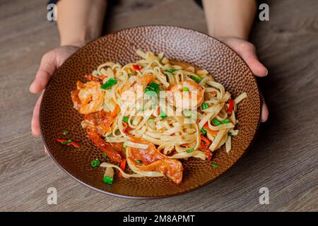 Foto ritagliata di una donna che tiene deliziosa pasta con gamberetti, verdi, pepe su un bel piatto marrone su un tavolo di legno. Foto Stock