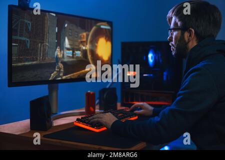 Giovane professionista caucasico che gioca in Online Cyber Sport Game a casa sua sul PC moderno. Uomo cyber sportivo partecipare tornei con la sua squadra. Vista laterale. Concetto di dipendenza da computer Foto Stock