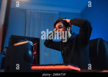 Ritratto di giovane giocatore di cyber sport maschile scioccato con il torneo online vincente o sparatutto video game, tiene le mani sulla sua testa, sentirsi uscito. Vista laterale, luce al neon, primo piano. Sport cyber professionale Foto Stock