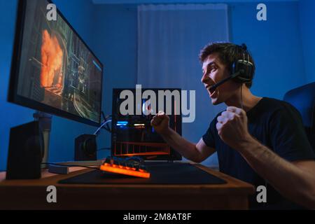 Cybersport giovane videogamer professionista felice di vincere il gioco, sentirsi uscito, mostrare sì gesto mano, celebra la vittoria nella competizione di gioco online. Vista laterale. Un ragazzo che gioca a un videogioco a casa sua in camera Foto Stock